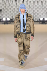 Male model wearing a beige patched suit with blue shirt and black tie, blue cap, walking the runway in a fashion show.