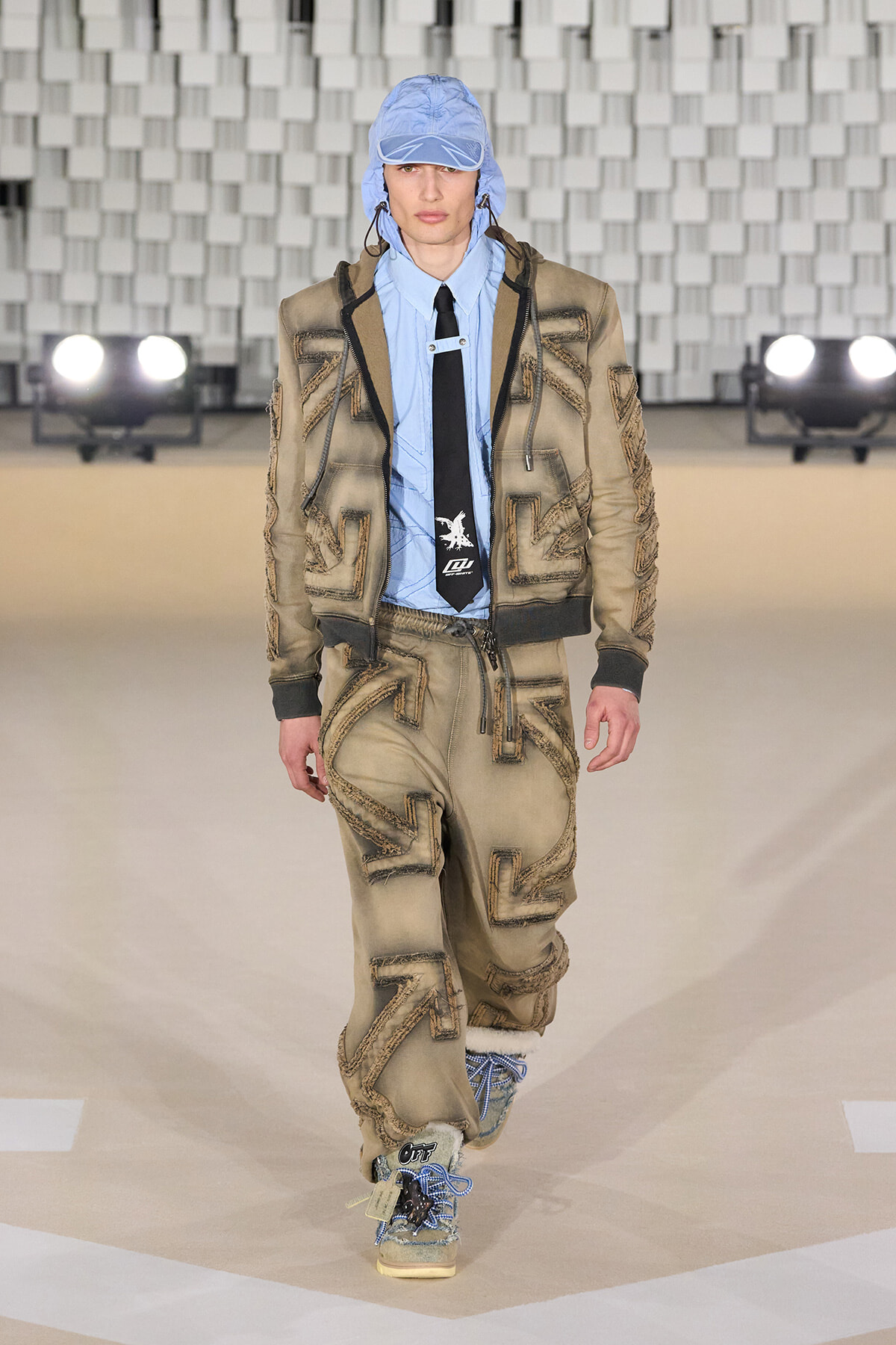 Male model wearing a beige patched suit with blue shirt and black tie, blue cap, walking the runway in a fashion show.