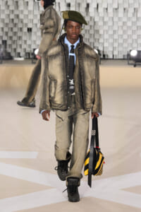 Male model walking a runway in a faded olive bomber jacket, light blue shirt, black tie, and green beret, carrying a yellow-and-black striped bag scroll.