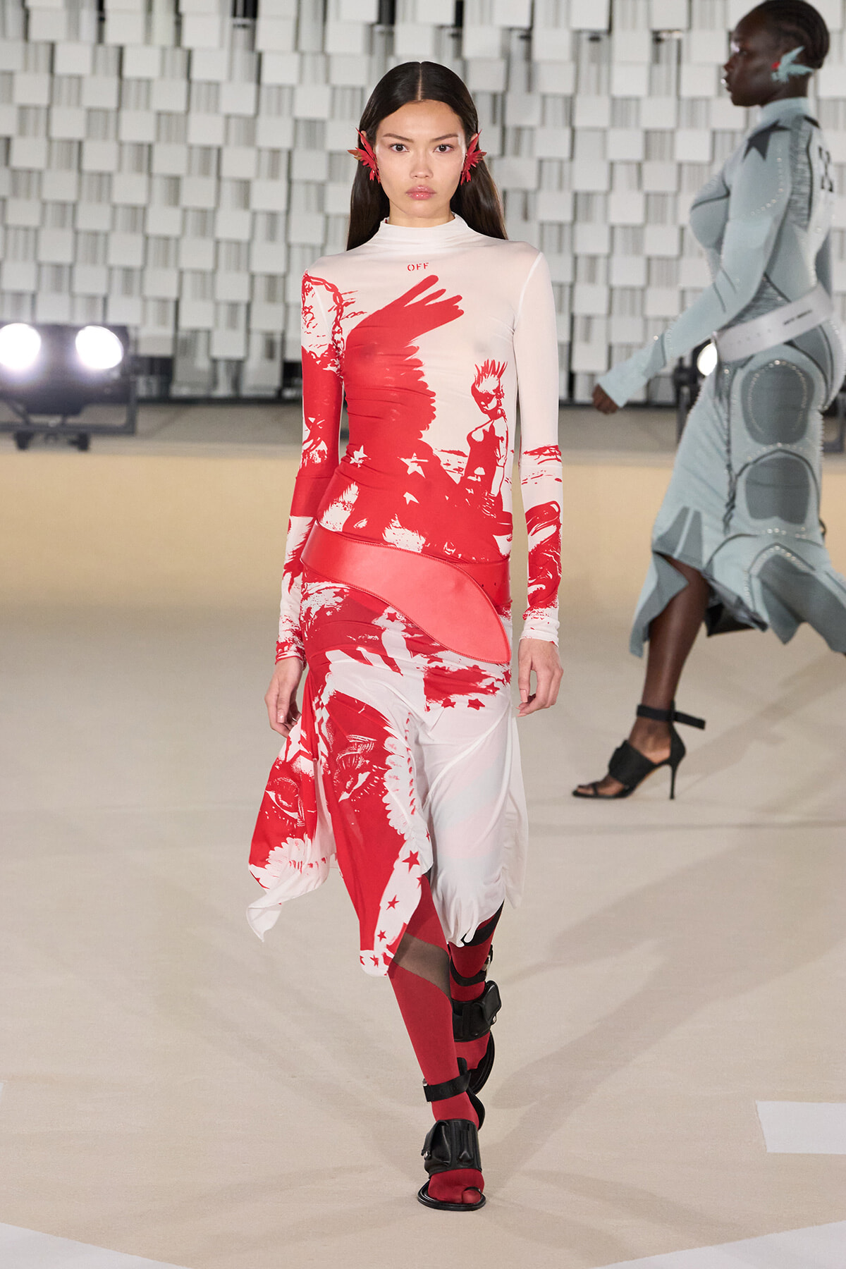 Model walks a runway in a white and red graphic knit dress with long sleeves, a pink belt-like wrap, red tights, and black strappy sandals.