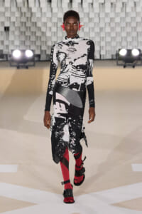 Model walks the fashion runway in a black-and-white abstract print outfit with red tights and chunky sandals, accented by red ear accessories on a bright stage backdrop.