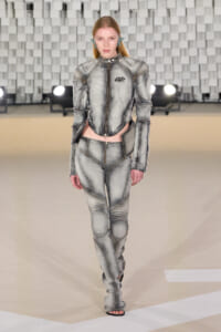 Model walks the runway in a metallic gray, armored-style jumpsuit with panel seams and exposed zippers on a fashion show catwalk.