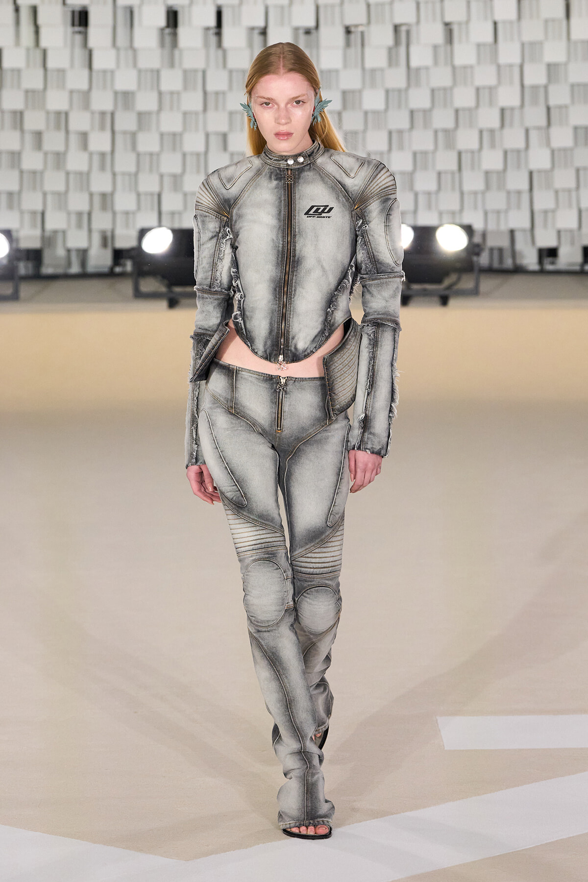 Model walks the runway in a metallic gray, armored-style jumpsuit with panel seams and exposed zippers on a fashion show catwalk.