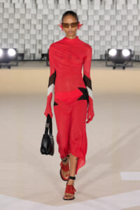 Model walks a fashion runway in a red, sculptural jumpsuit with black and white color-block accents, red sandals, brown sunglasses, and a black handbag.