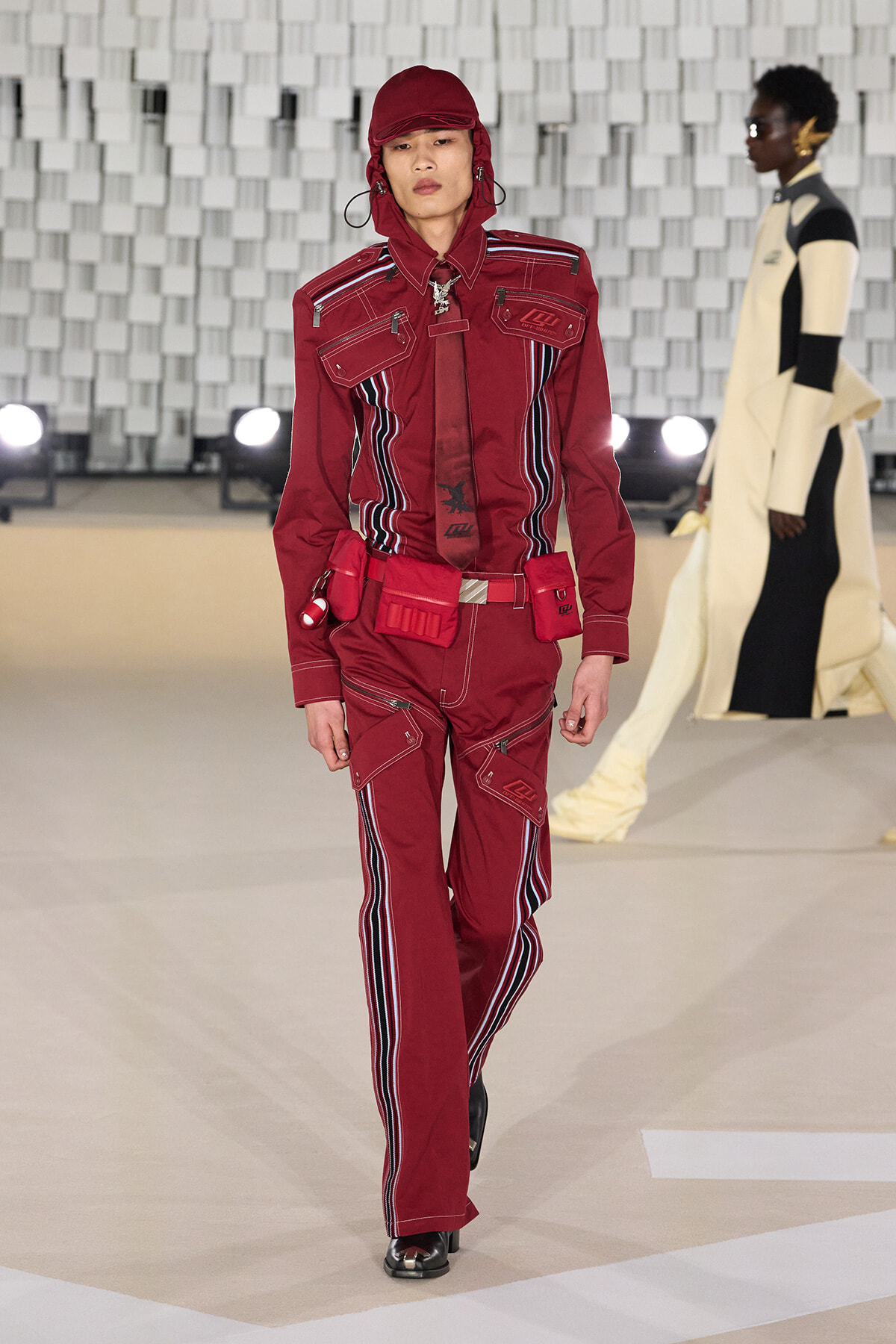 Model walking a fashion runway in a red utility jumpsuit with hood and striped side panels.