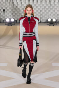 Model walking a runway in a red, gray, and black color-block varsity-inspired outfit, carrying a black bag.