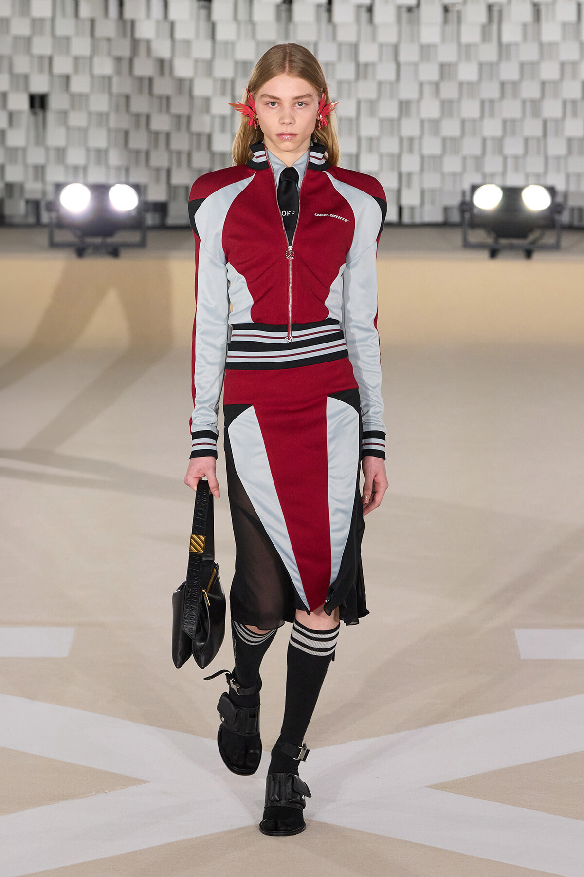 Model walking a runway in a red, gray, and black color-block varsity-inspired outfit, carrying a black bag.