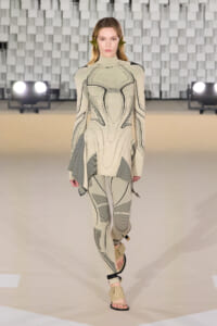 Model walks the runway in a beige geometric-print bodysuit with an asymmetric tunic and tan sandals.