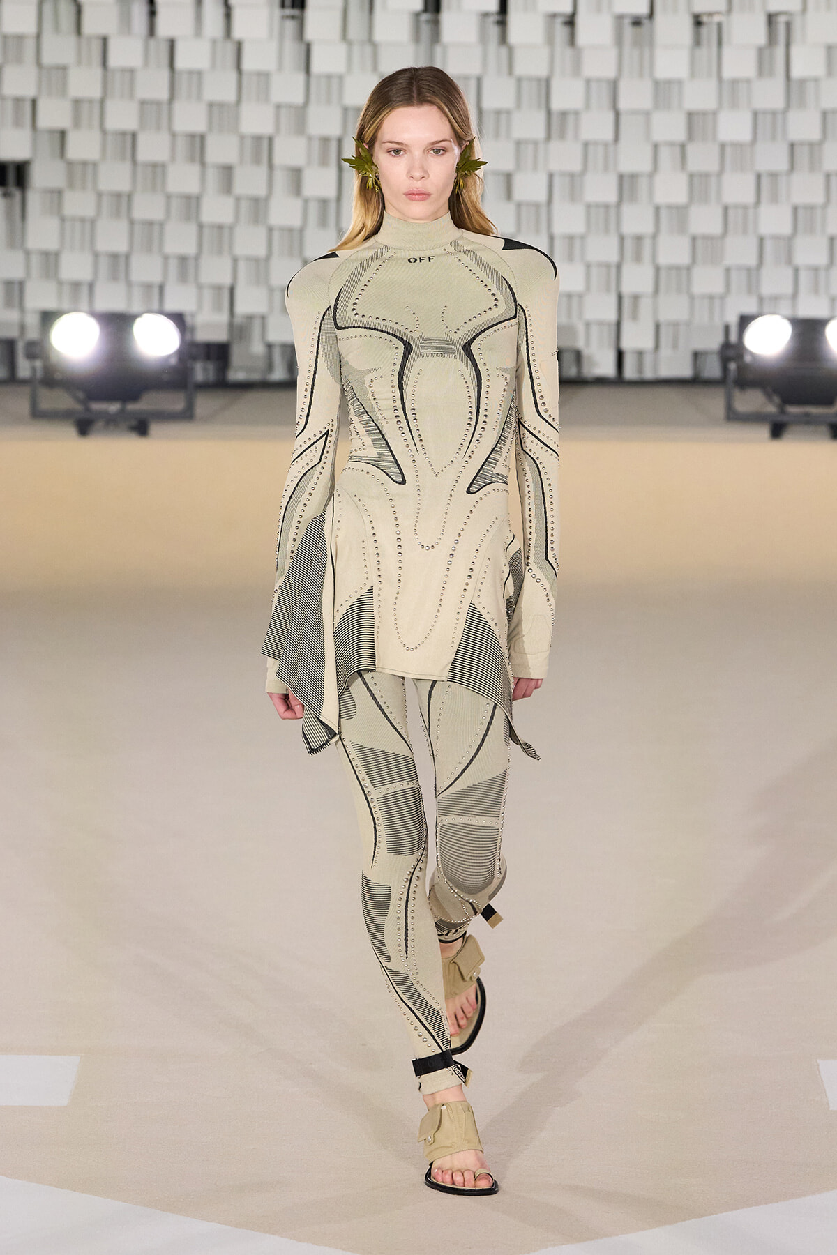 Model walks the runway in a beige geometric-print bodysuit with an asymmetric tunic and tan sandals.