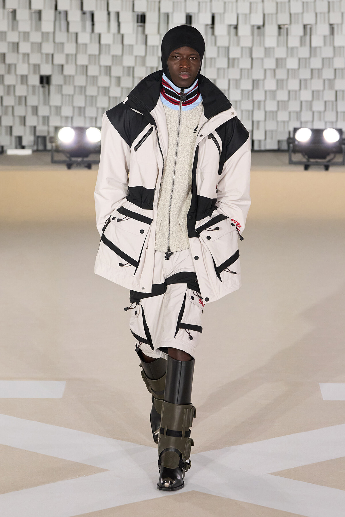 Male model walks a fashion runway in a white and black oversized parka with multiple pockets, paired with matching shorts and knee-high boots.