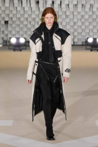 Model walks the runway in a long black-and-white color-blocked coat with multiple zippers and a black outfit beneath.
