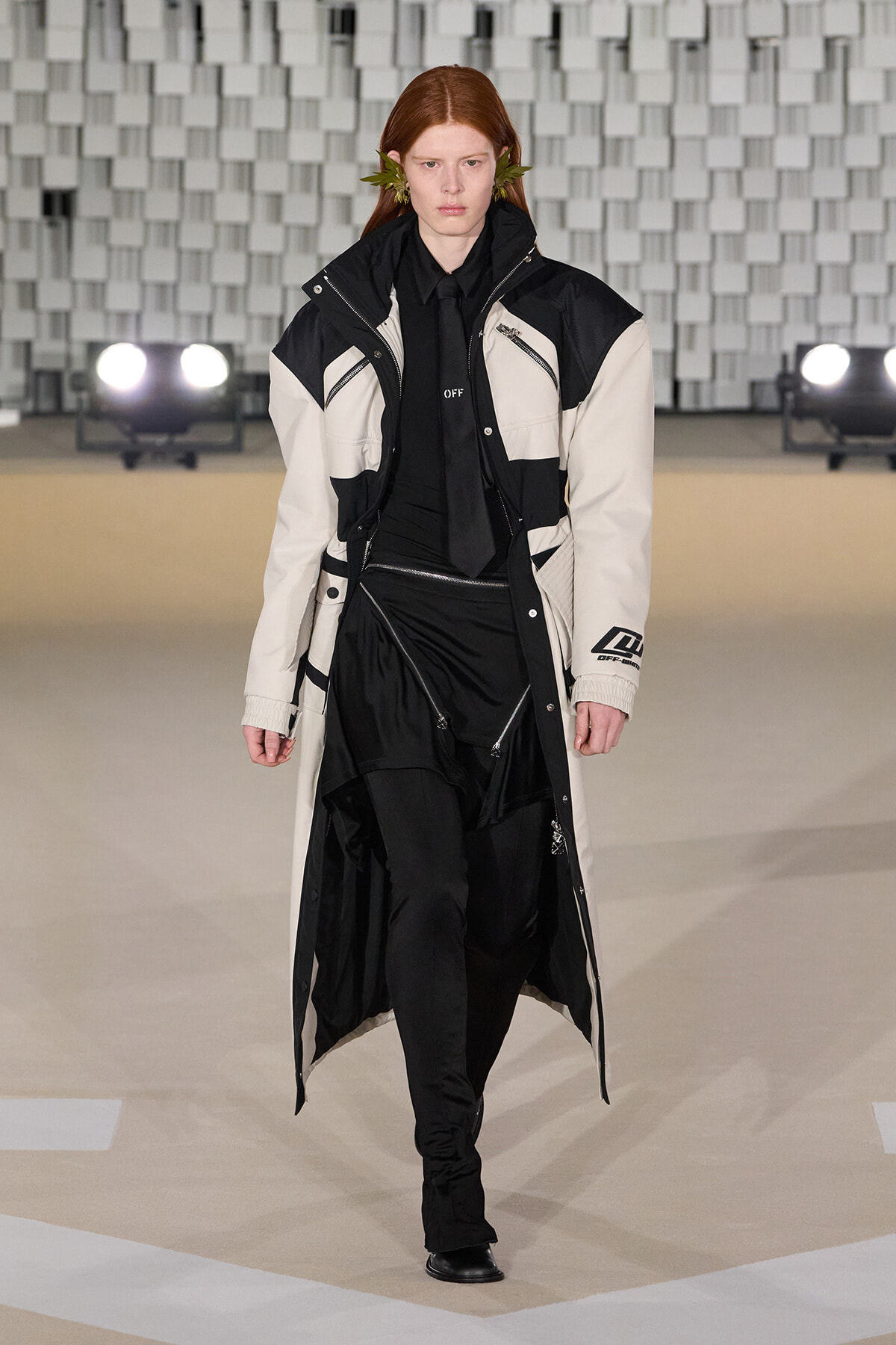 Model walks the runway in a long black-and-white color-blocked coat with multiple zippers and a black outfit beneath.