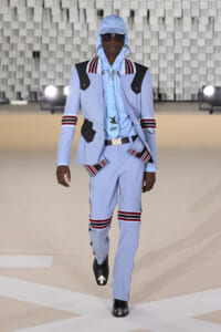 Male model wearing a light blue tailored suit with black-and-red striped accents on a runway, accessorized with a matching cap, sunglasses, and white belt.