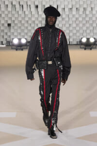 Male model walks a fashion runway wearing a black utility jumpsuit with red piping and white star details, accessorized with a belt of pouches and a black beret-style hat.