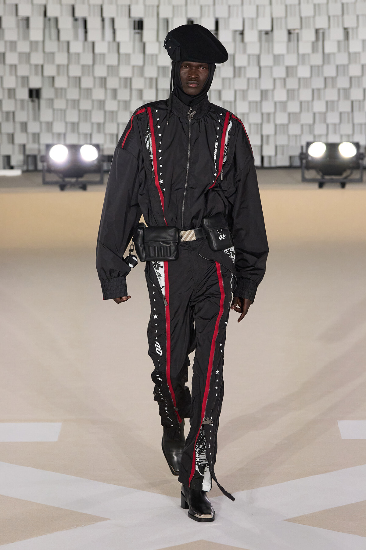 Male model walks a fashion runway wearing a black utility jumpsuit with red piping and white star details, accessorized with a belt of pouches and a black beret-style hat.