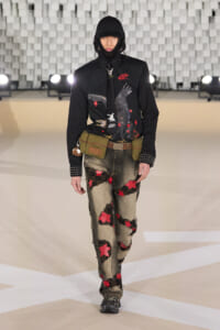 Model walking a runway in a black hooded jacket with an eagle graphic, olive utility belt, and patchwork jeans with red stars and patches.
