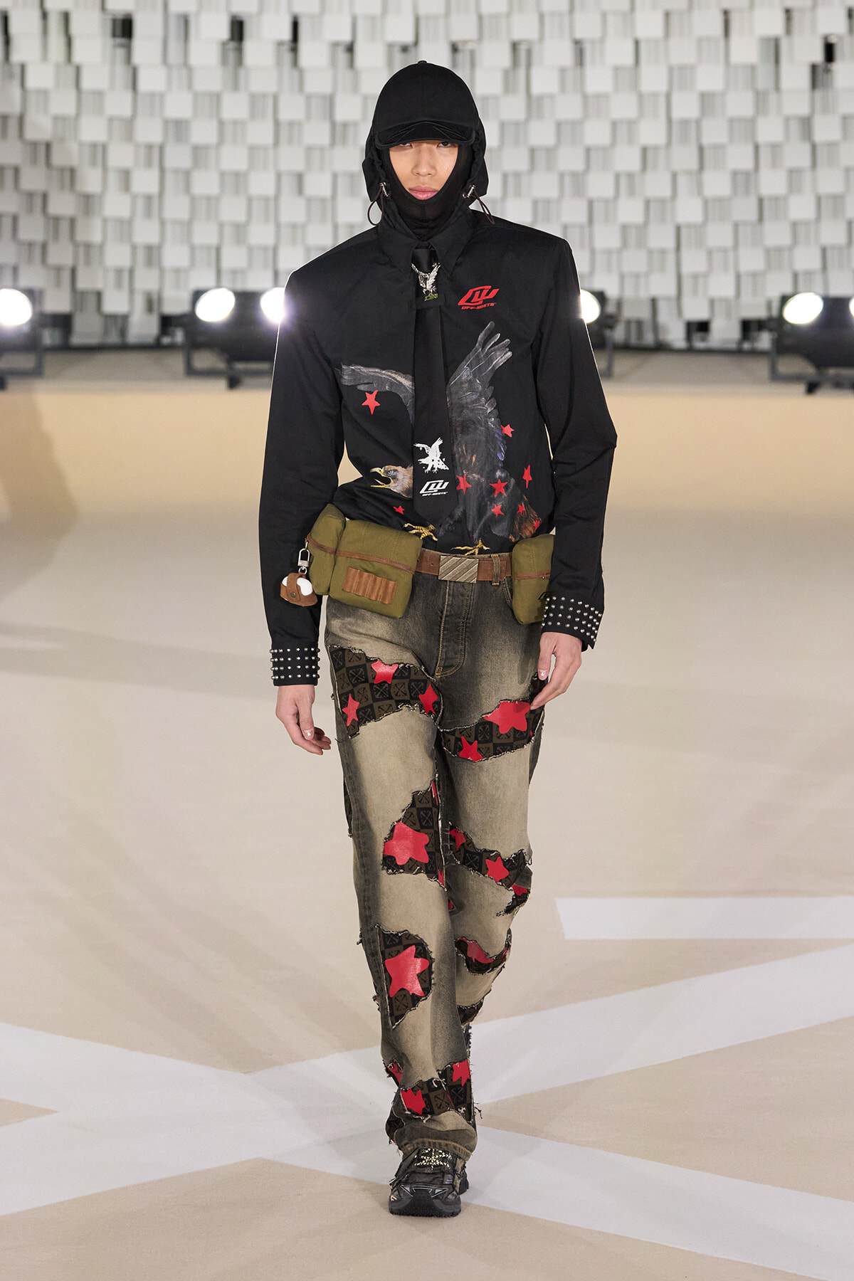 Model walking a runway in a black hooded jacket with an eagle graphic, olive utility belt, and patchwork jeans with red stars and patches.