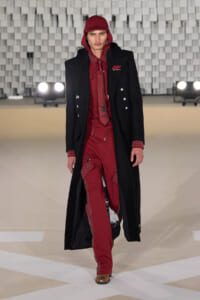 Male model walking a runway in a red studded suit beneath a long black coat and red cap, showcasing a high-fashion look on a neutral stage backdrop.