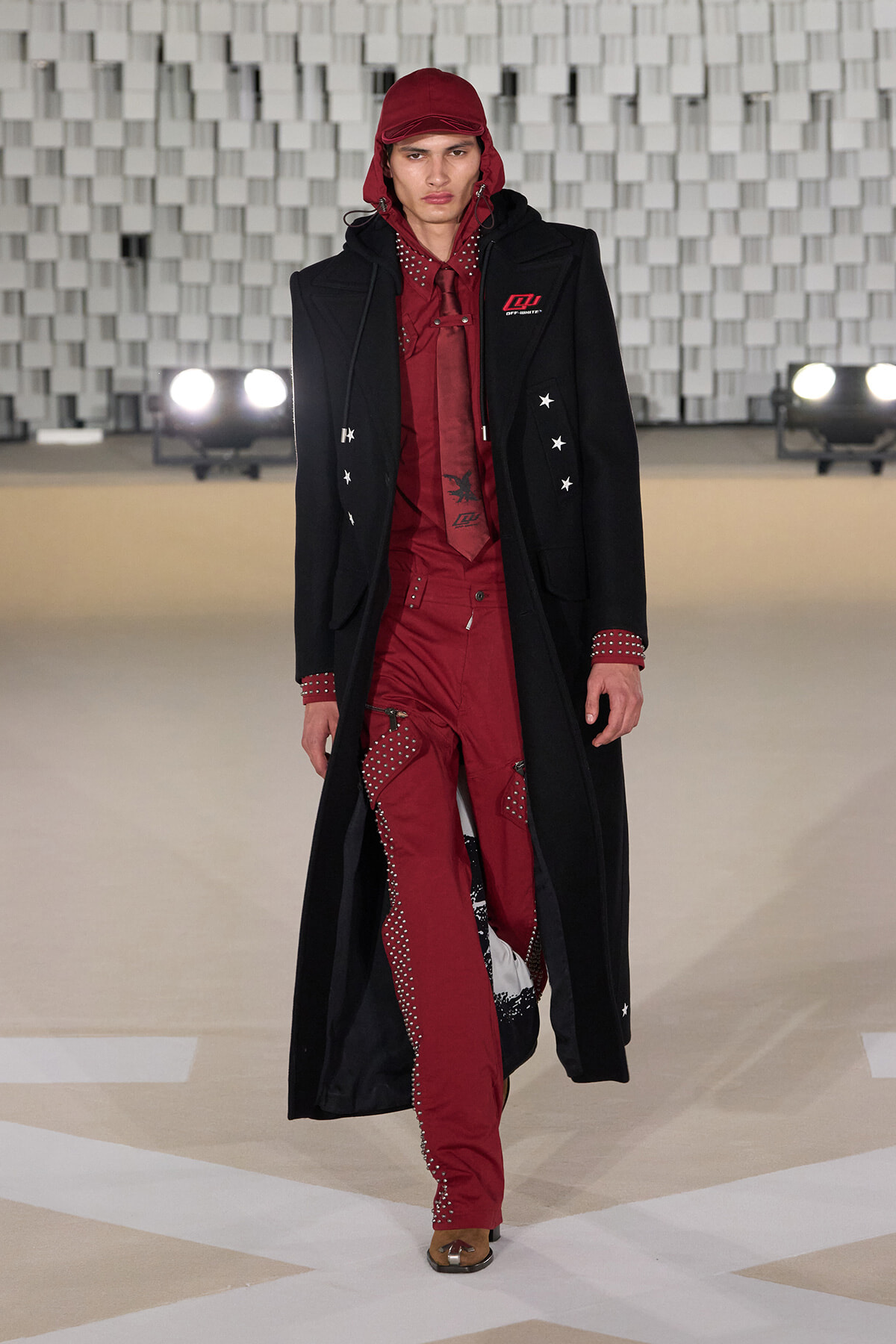 Male model walking a runway in a red studded suit beneath a long black coat and red cap, showcasing a high-fashion look on a neutral stage backdrop.