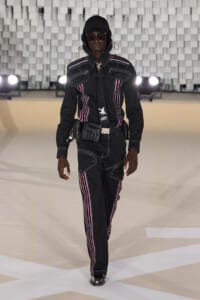 Model walking a fashion runway in a black utilitarian suit with red, white, and blue piping, hood, sunglasses, and utility belt accessories.
