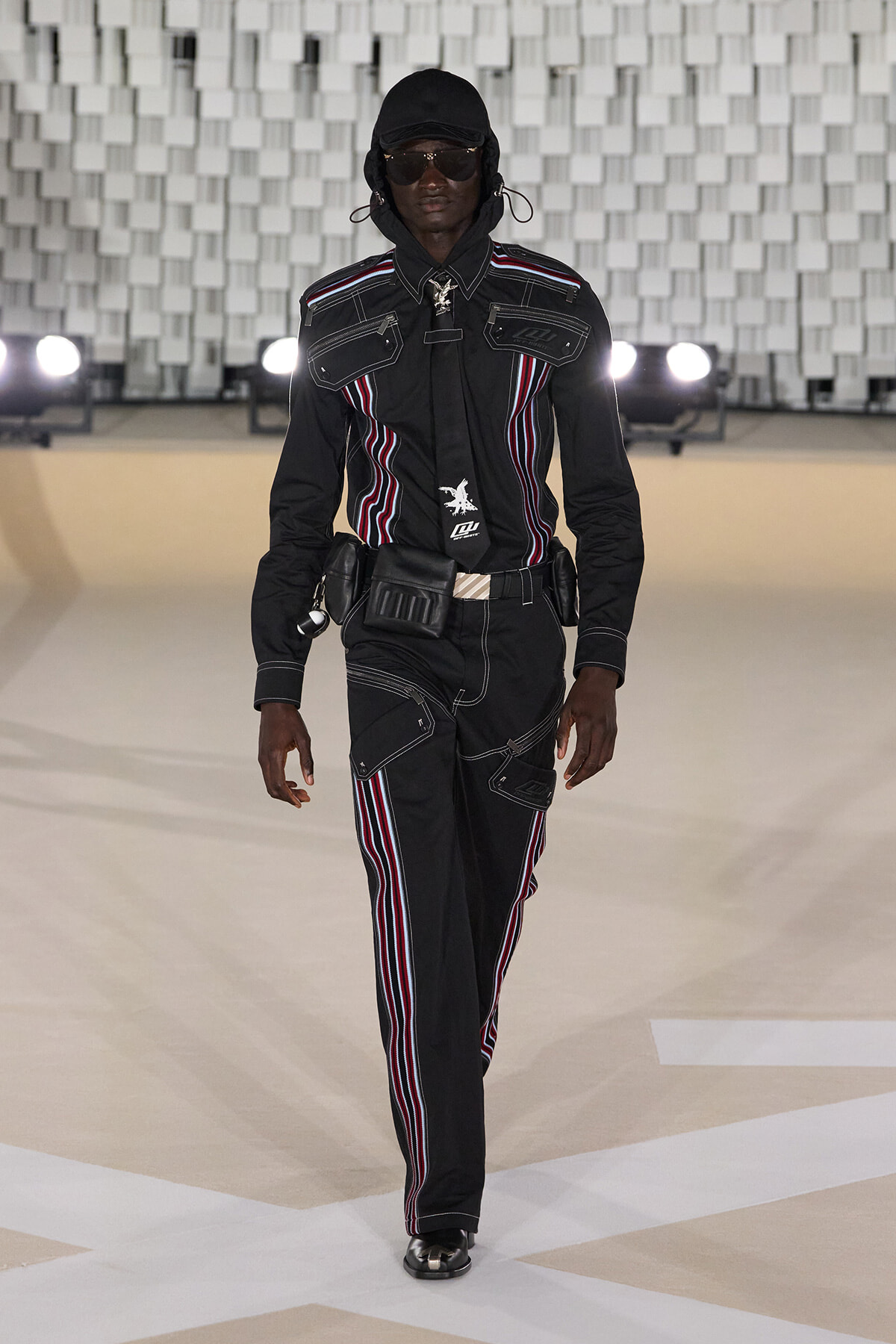 Model walking a fashion runway in a black utilitarian suit with red, white, and blue piping, hood, sunglasses, and utility belt accessories.