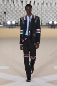 Male model walking a runway in a black suit with red and white striped accents, light blue shirt, and black boots against a geometric backdrop.