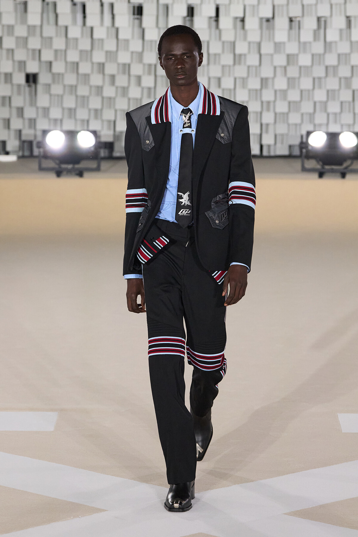 Male model walking a runway in a black suit with red and white striped accents, light blue shirt, and black boots against a geometric backdrop.