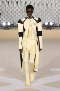 Model on a runway wearing a cream and black color-blocked long coat with matching pale yellow outfit and knee-high boots, sunglasses, and gold ear accessories.