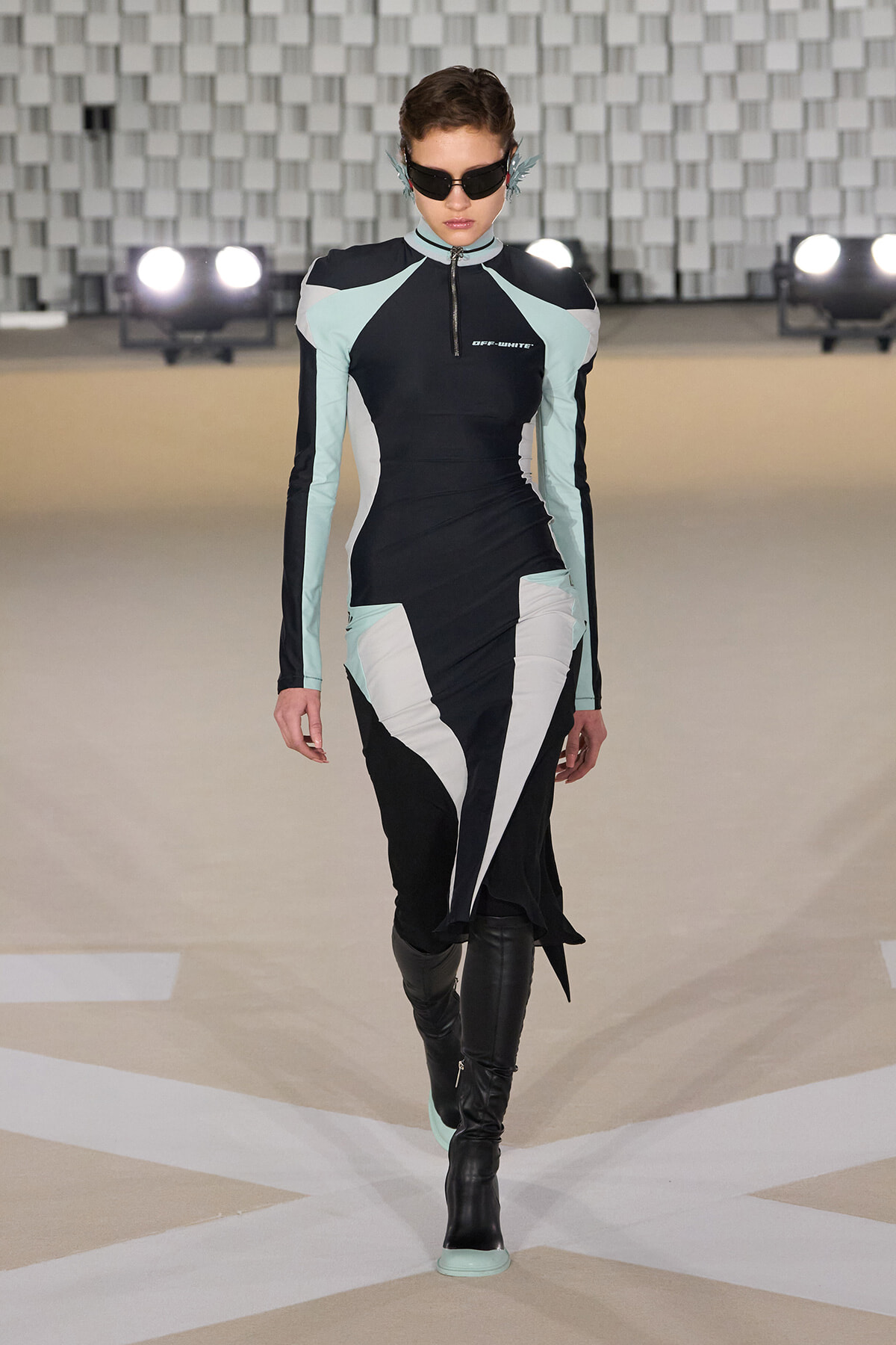 Model walking a runway in a color-block, form-fitting black dress with mint and white panels, paired with black knee-high boots and futuristic sunglasses.