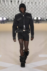 Model walks the runway in a black studded outfit with a hood, cap, and oversized belt pouches; knee-high, strapped black boots add a futuristic vibe.