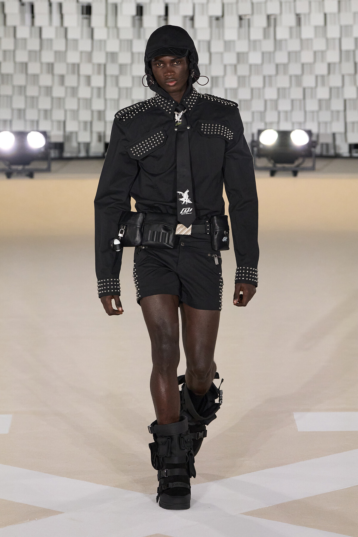 Model walks the runway in a black studded outfit with a hood, cap, and oversized belt pouches; knee-high, strapped black boots add a futuristic vibe.