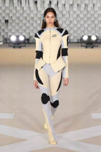 Model walking the runway in a cream, gray, and black color-blocked futuristic bodysuit with armor-like panels and matching boots.