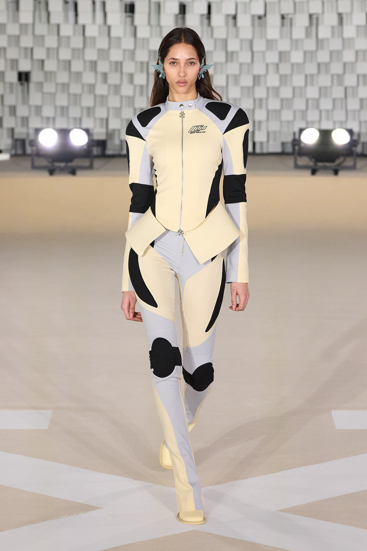 Model walking the runway in a cream, gray, and black color-blocked futuristic bodysuit with armor-like panels and matching boots.