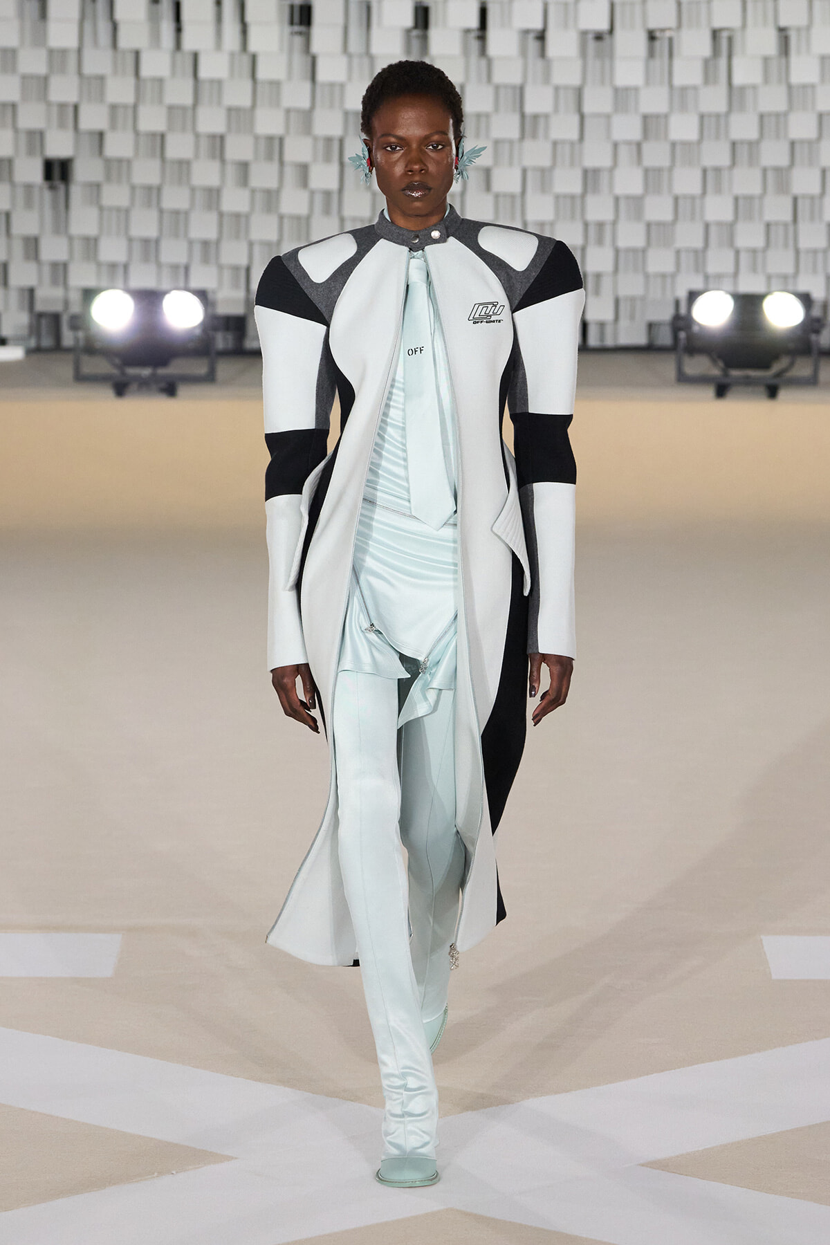 Model walks a fashion runway in a white and black structured coat over a pale blue, asymmetric suit with long sleeves and high collar, set against a geometric backdrop with spotlights.