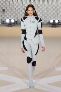 Model walking a fashion show in a futuristic mint-green and black space-inspired bodysuit with matching boots; geometric panels and zipped front.
