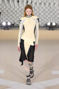 Female model walks a runway in a cream and gray geometric top with an asymmetrical black-and-cream skirt and sheer panels, futuristic footwear, on a fashion-show stage with grid backdrop.