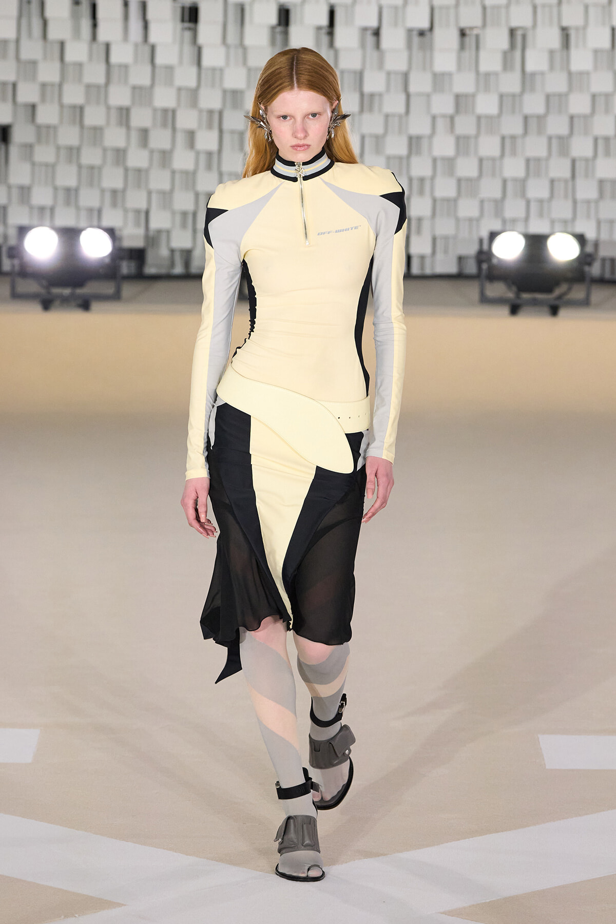 Female model walks a runway in a cream and gray geometric top with an asymmetrical black-and-cream skirt and sheer panels, futuristic footwear, on a fashion-show stage with grid backdrop.