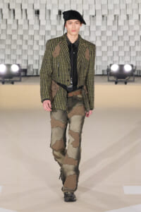 Male model on a runway wearing a green tweed blazer with brown patches, black shirt, patchwork jeans, and a black beret.