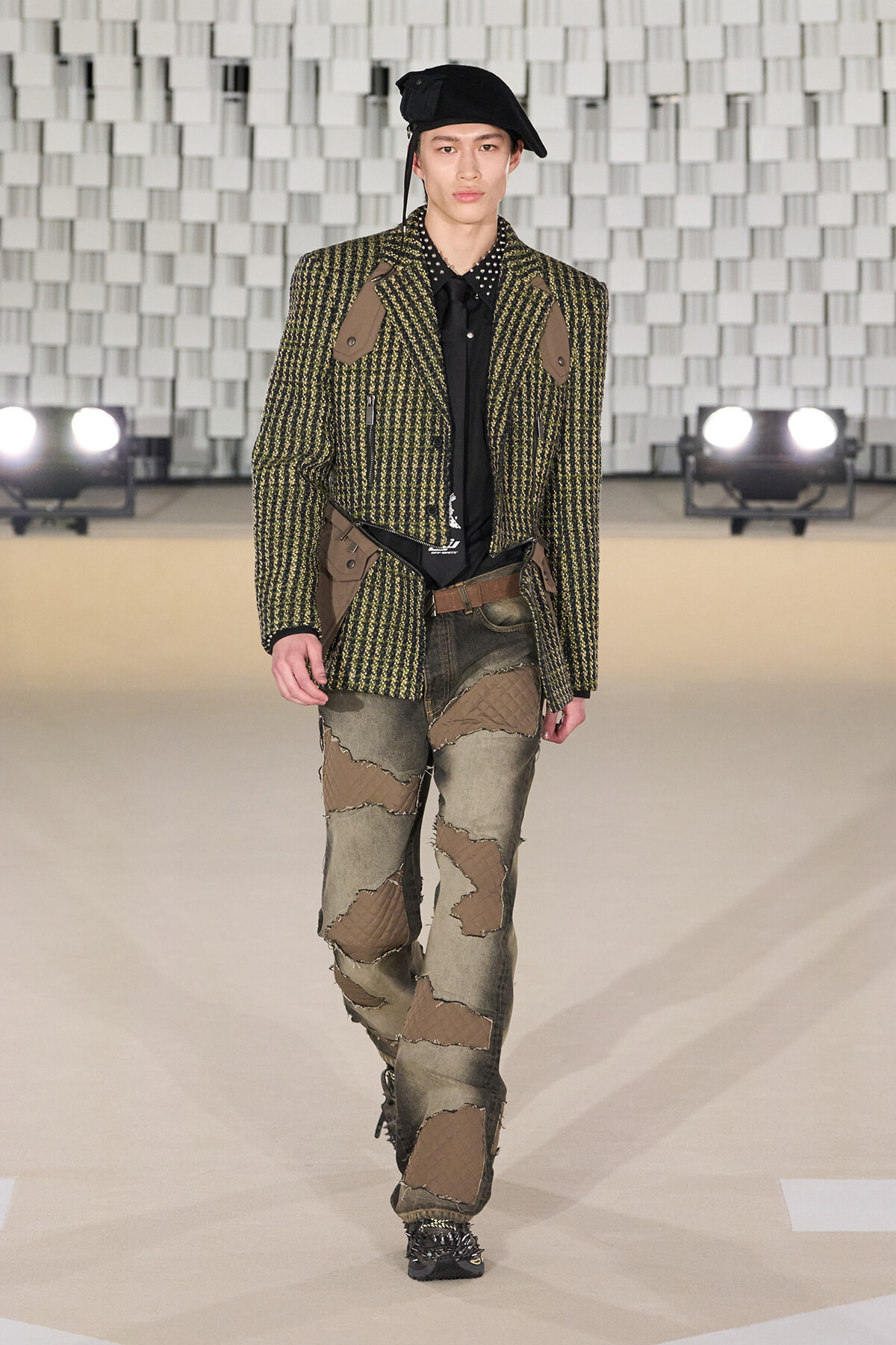 Male model on a runway wearing a green tweed blazer with brown patches, black shirt, patchwork jeans, and a black beret.