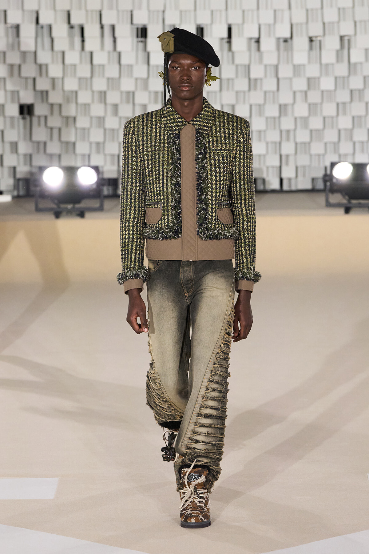 Male model on a runway wearing a green tweed jacket with fringe trim, beige accents, and distressed jeans with camo boots, black beret.