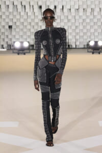 Model walking the runway in a black-and-white geometric jumpsuit with cutouts at the waist, high collar, sunglasses, and statement earrings.