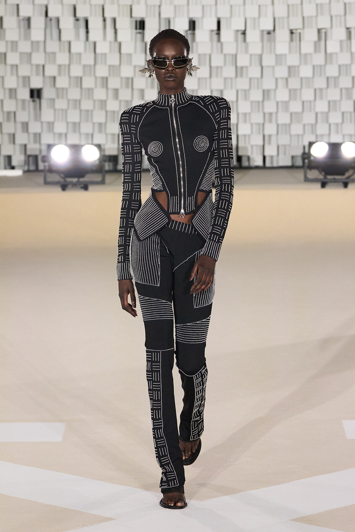 Model walking the runway in a black-and-white geometric jumpsuit with cutouts at the waist, high collar, sunglasses, and statement earrings.