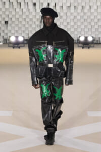Model walking a runway in a glossy black jumpsuit with neon-green bird graphics, paired with a black beret and boots.