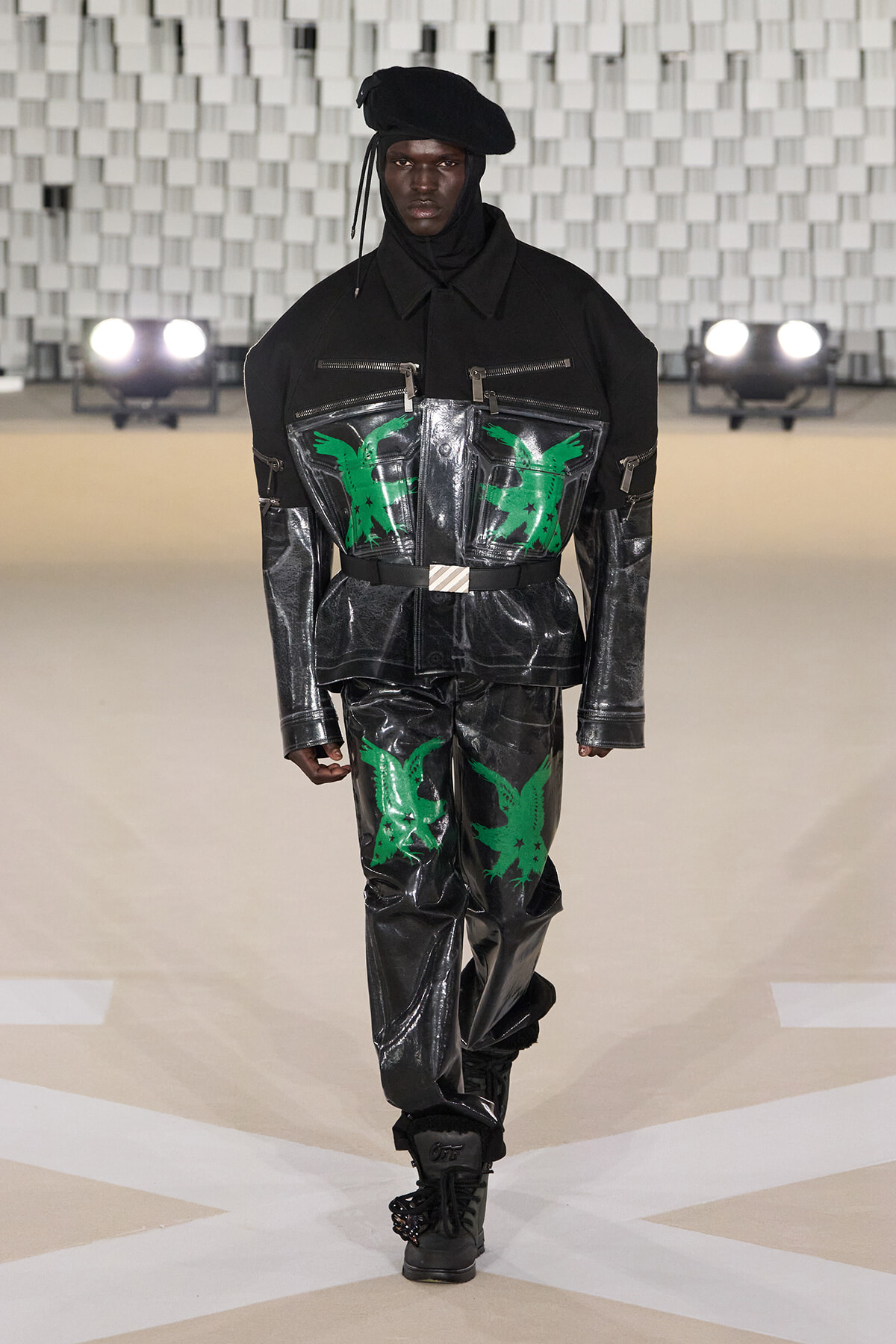 Model walking a runway in a glossy black jumpsuit with neon-green bird graphics, paired with a black beret and boots.
