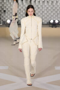 Model walks the runway in a cream textured suit with fringe, high collar, and a silver zip, against a geometric backdrop.