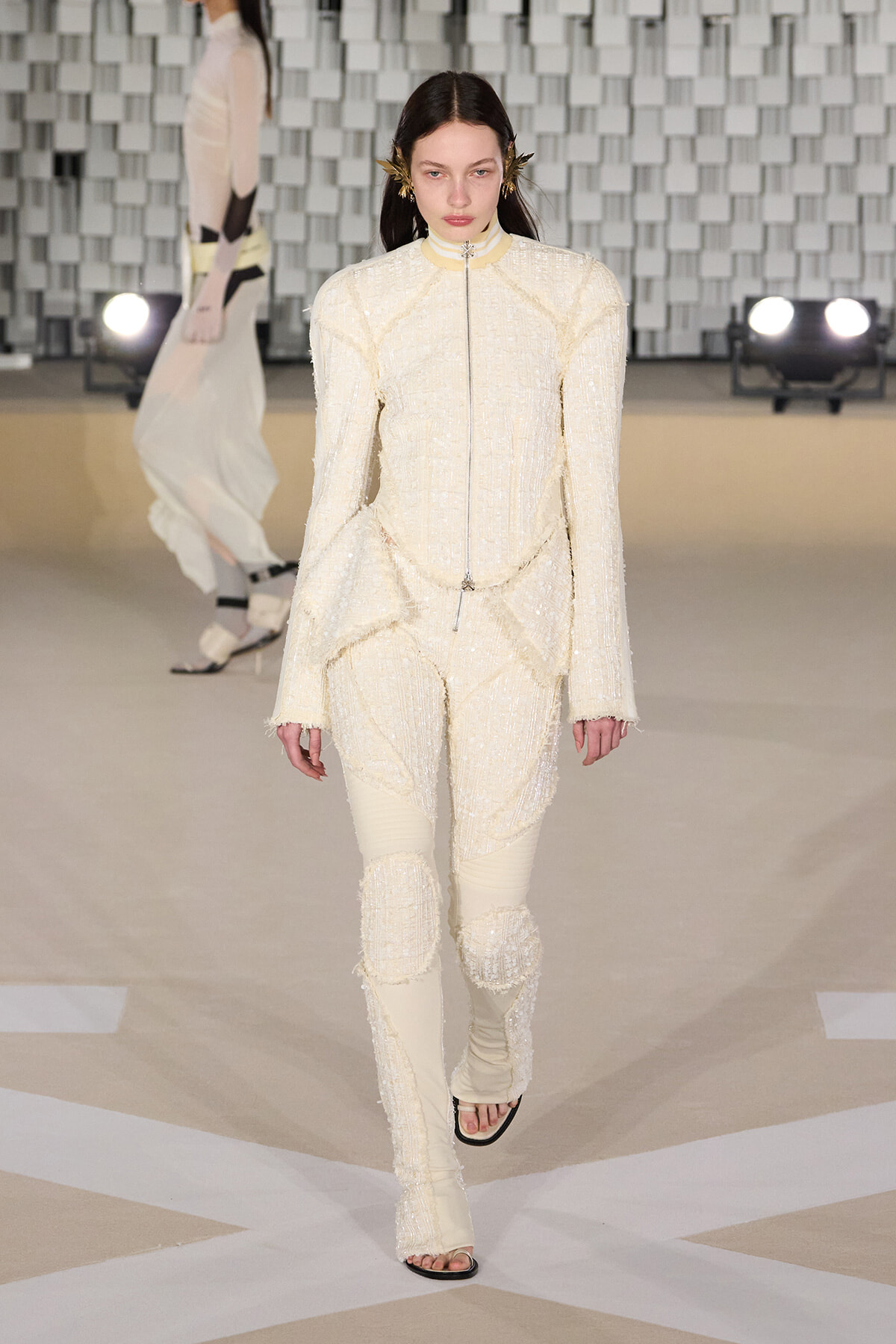 Model walks the runway in a cream textured suit with fringe, high collar, and a silver zip, against a geometric backdrop.