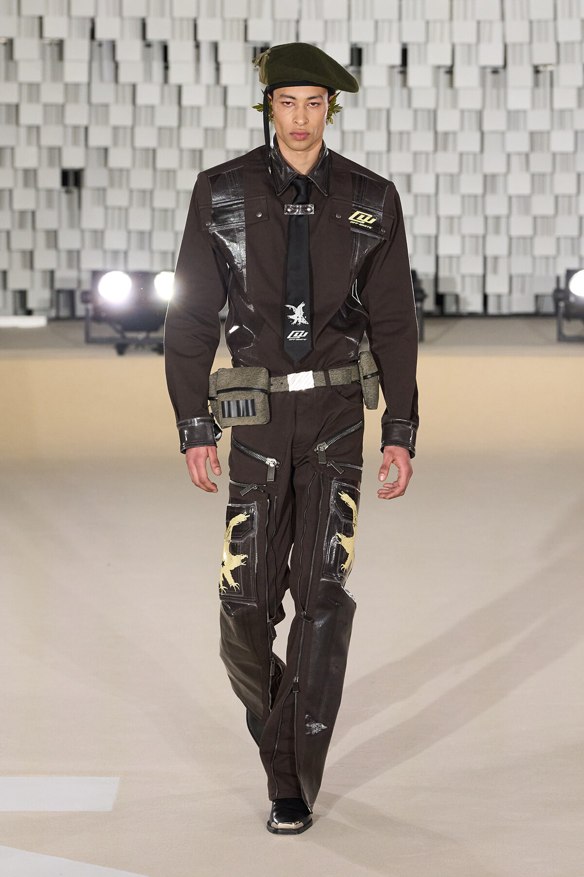 Male model walking a runway in a dark brown utility jumpsuit with patches, a black tie, and an olive beret on a fashion stage.