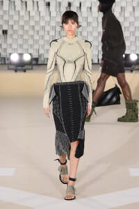 Model on a runway wearing a beige sculpted top with studded detailing and a black geometric-patterned midi skirt, sandals visible.