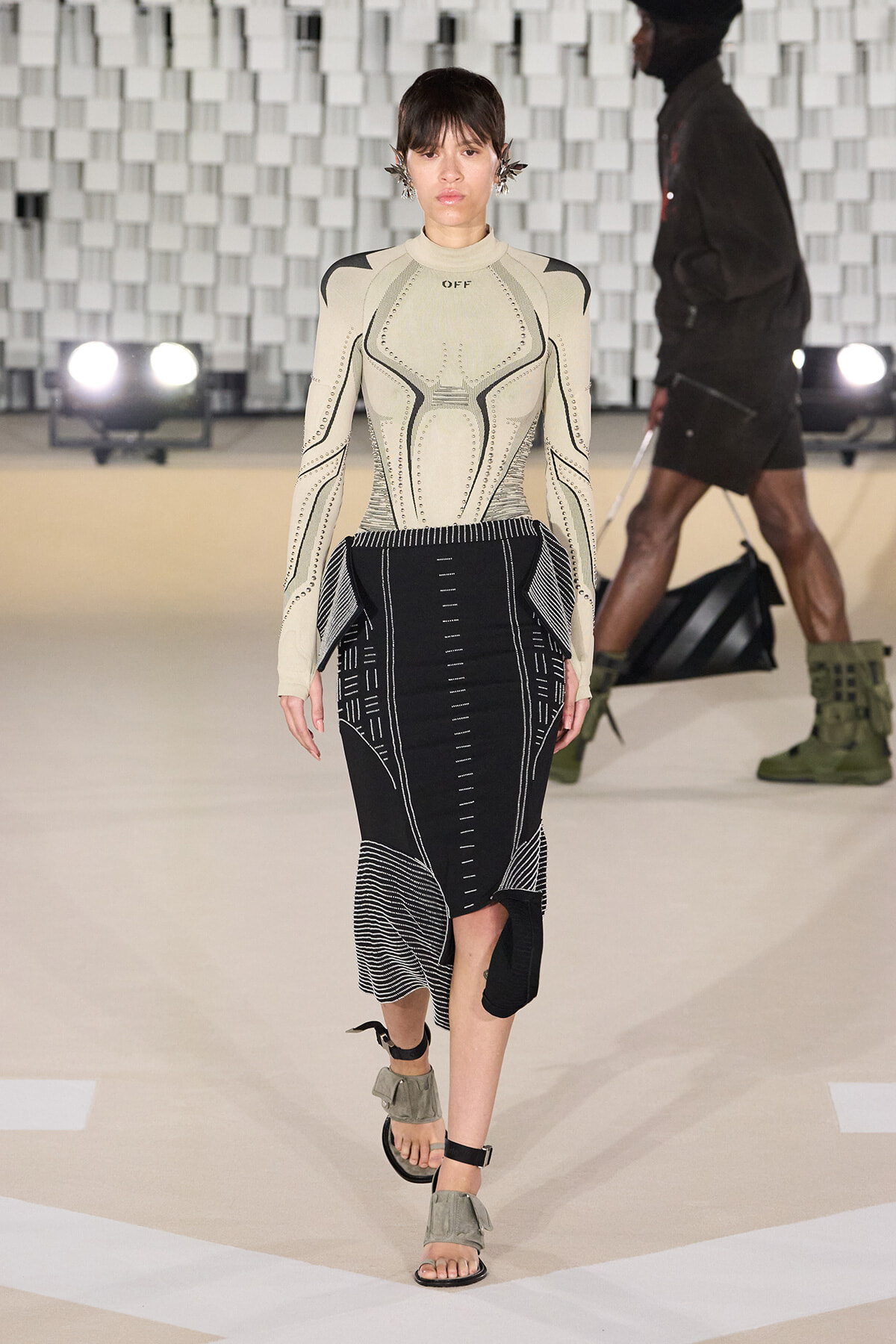 Model on a runway wearing a beige sculpted top with studded detailing and a black geometric-patterned midi skirt, sandals visible.
