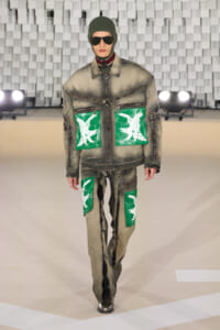 Male model on a runway wearing a distressed gray jacket and pants with bright green patch pockets featuring white bird graphics; sunglasses and a green balaclava.